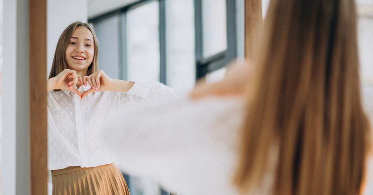 Woman smiling in the mirror while placing her hands in the shape of a heart.